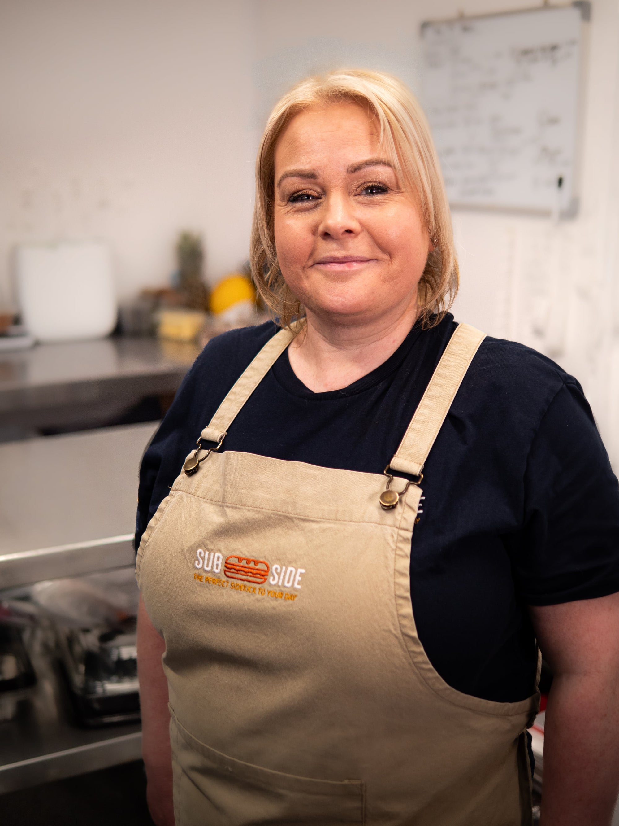 A woman wearing a beige "Sub Side" apron stands in a commercial kitchen, looking at the camera and smiling.