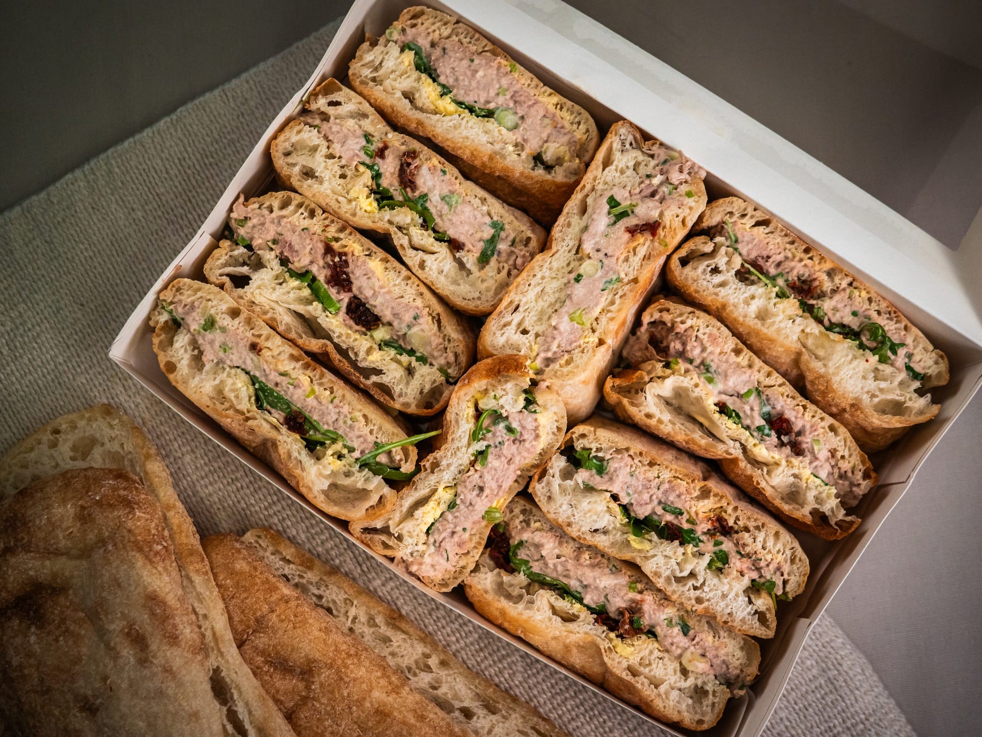 The Sub Side Mixed Sub Platter - 2 Full Boxes (serves 10) features assorted halved signature subs with ground meat, greens, and sauce, presented beside pieces of bread on a cloth surface.