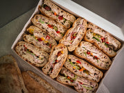 The Sub Side Roasted Chicken Crunch box features assorted ciabatta sandwiches with sliced chicken breast, mixed peppers, hot red onion mayo, various meats, veggies, spreads, and extra bread pieces on the side.