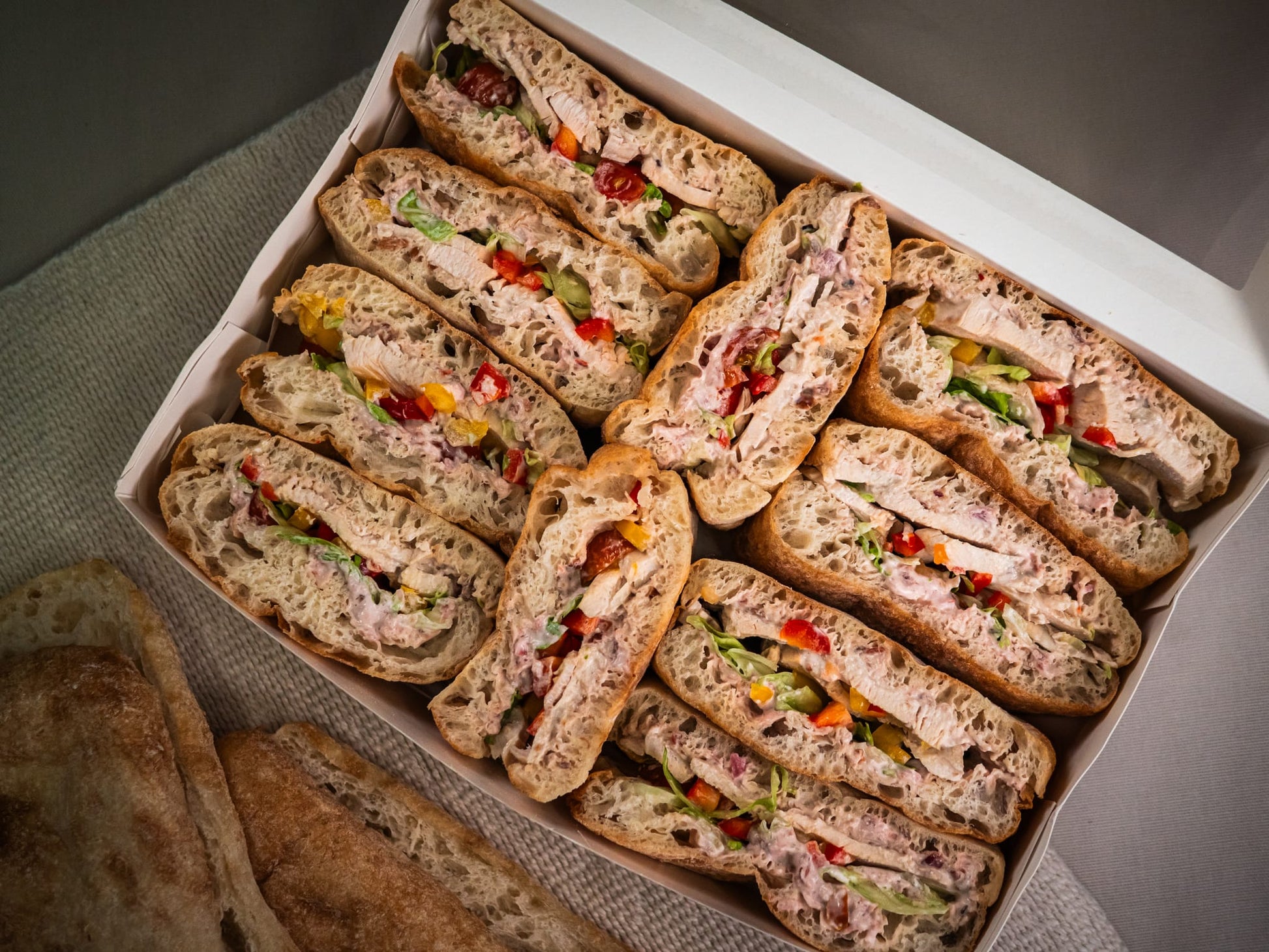 The Sub Side Roasted Chicken Crunch box features assorted ciabatta sandwiches with sliced chicken breast, mixed peppers, hot red onion mayo, various meats, veggies, spreads, and extra bread pieces on the side.