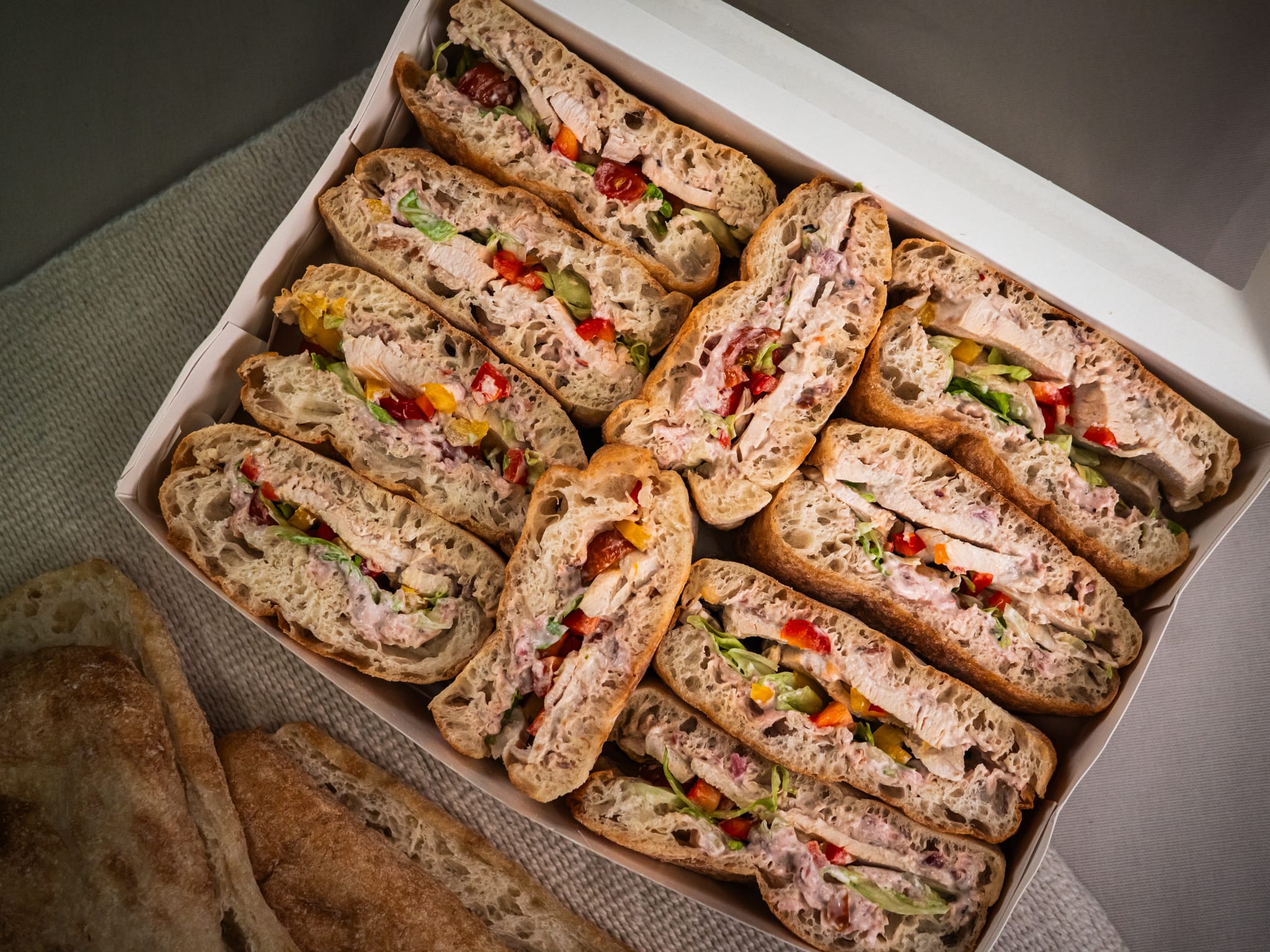 The Sub Side Roasted Chicken Crunch box features assorted ciabatta sandwiches with sliced chicken breast, mixed peppers, hot red onion mayo, various meats, veggies, spreads, and extra bread pieces on the side.