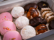 The Baker’s Box (15 pieces) by Sub Side features assorted pastries: pink glazed cookies with sprinkles, coconut-covered treats, chocolate eclairs, iced jam biscuits, chocolate pastries, and coconut bars.