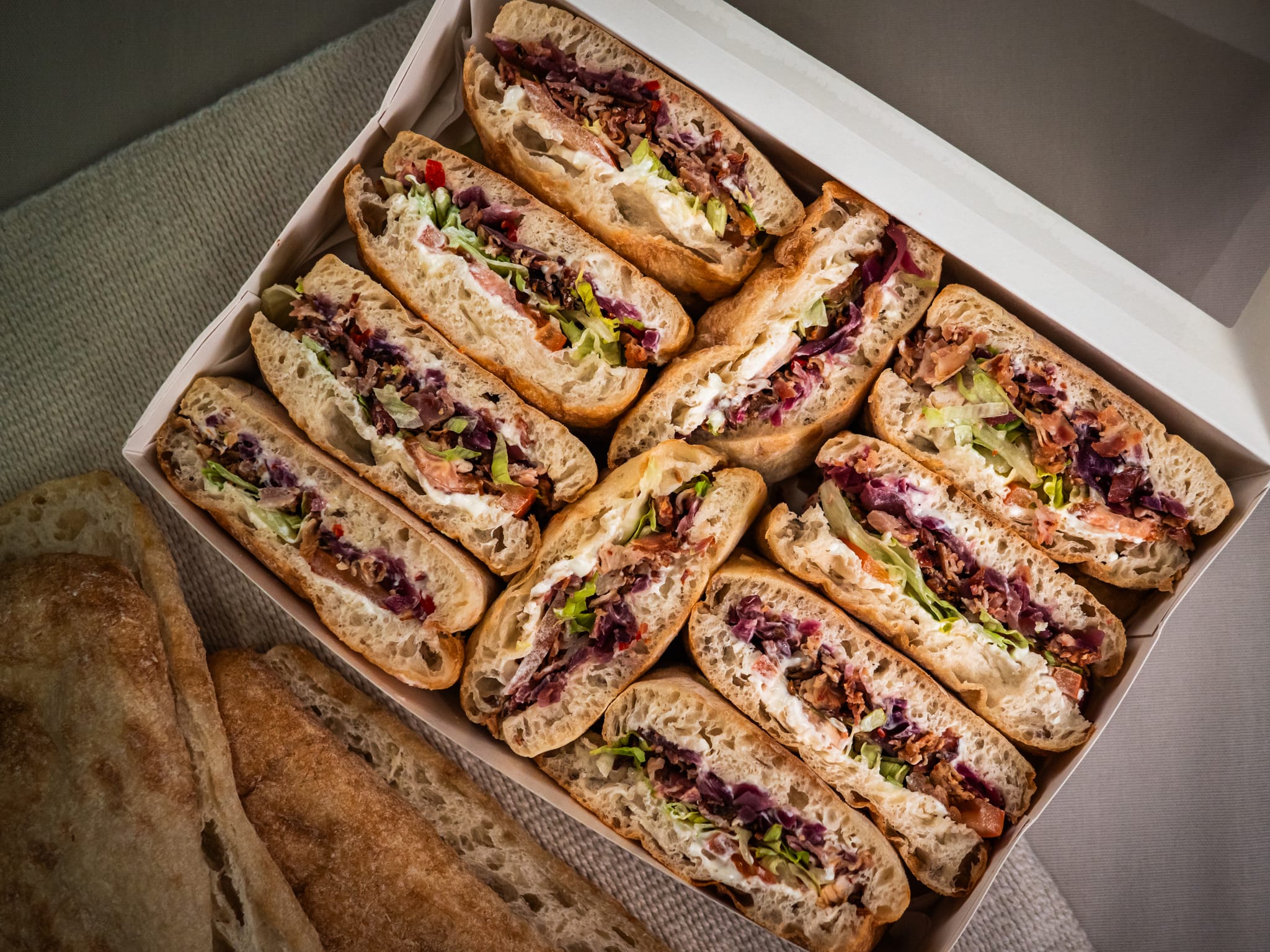 The Sub Side Mixed Sub Platter - 2 Full Boxes (serves 10) features signature and vegetarian subs with lettuce, deli meats, and veggies, arranged neatly in rows. Slices of bread are served alongside the platter.