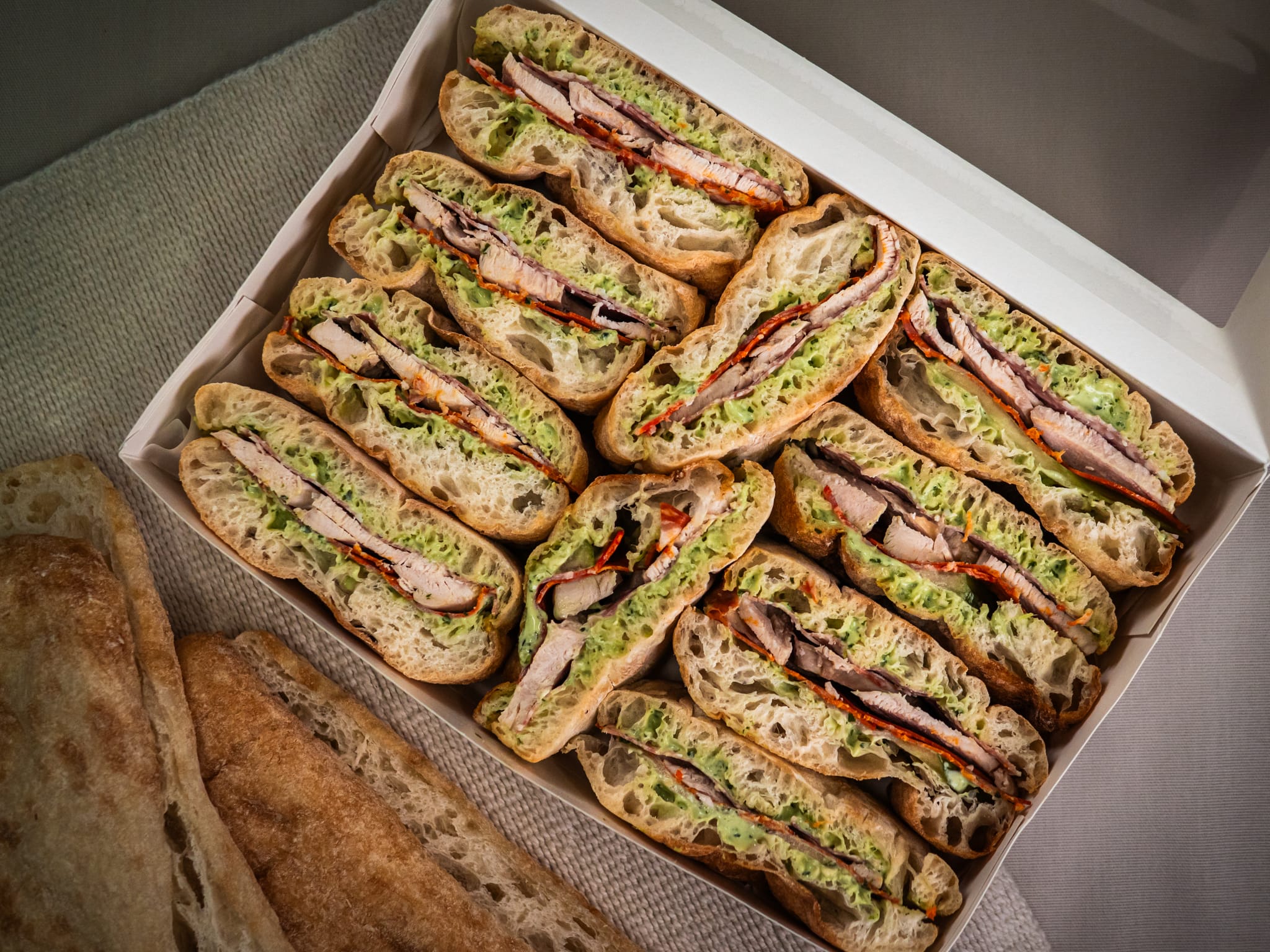 The Sub Side Hearty Sub Side features ciabatta sandwiches with roasted chicken, lettuce, and roasted red peppers in creamy pesto mayo, arranged neatly and served with extra bread.