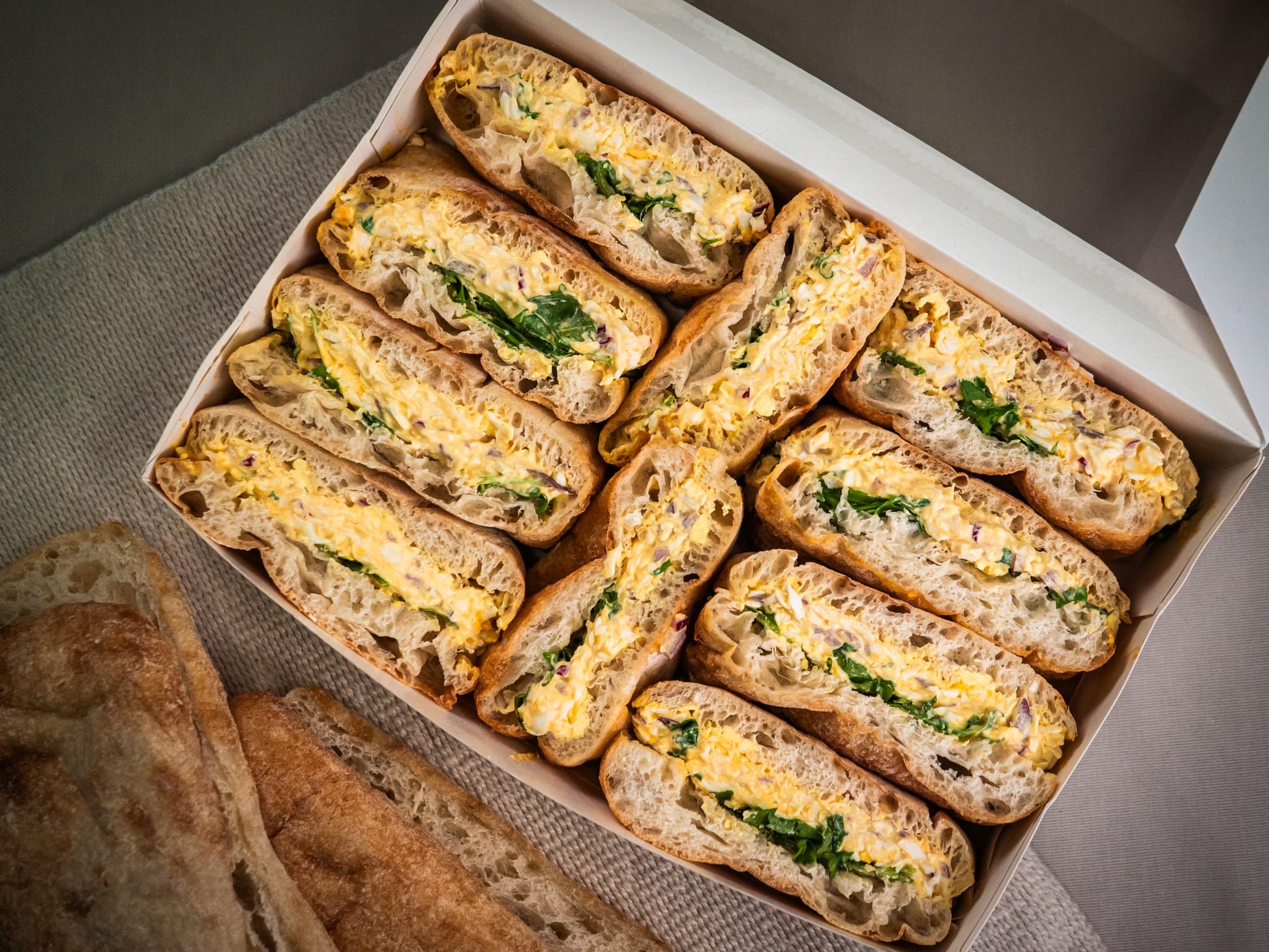 A Sub Side Sunshine Stack box with ten sandwiches filled with creamy Dijon egg salad and fresh rocket, served on a cloth next to slices of bread.