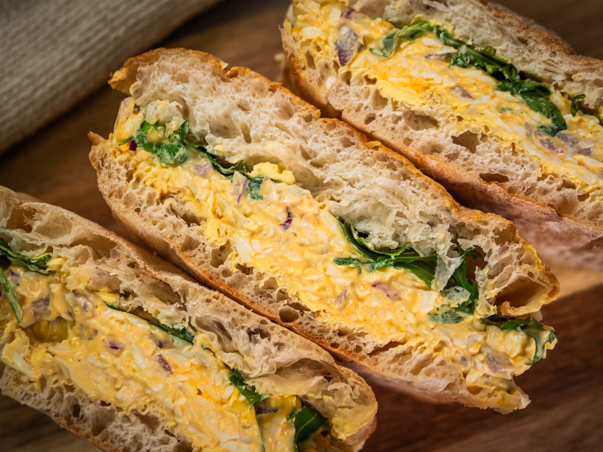 Three halves of Sub Side's Sunshine Stack, a sandwich filled with creamy Dijon egg salad, fresh rocket, and chopped vegetables, are arranged on a wooden surface.