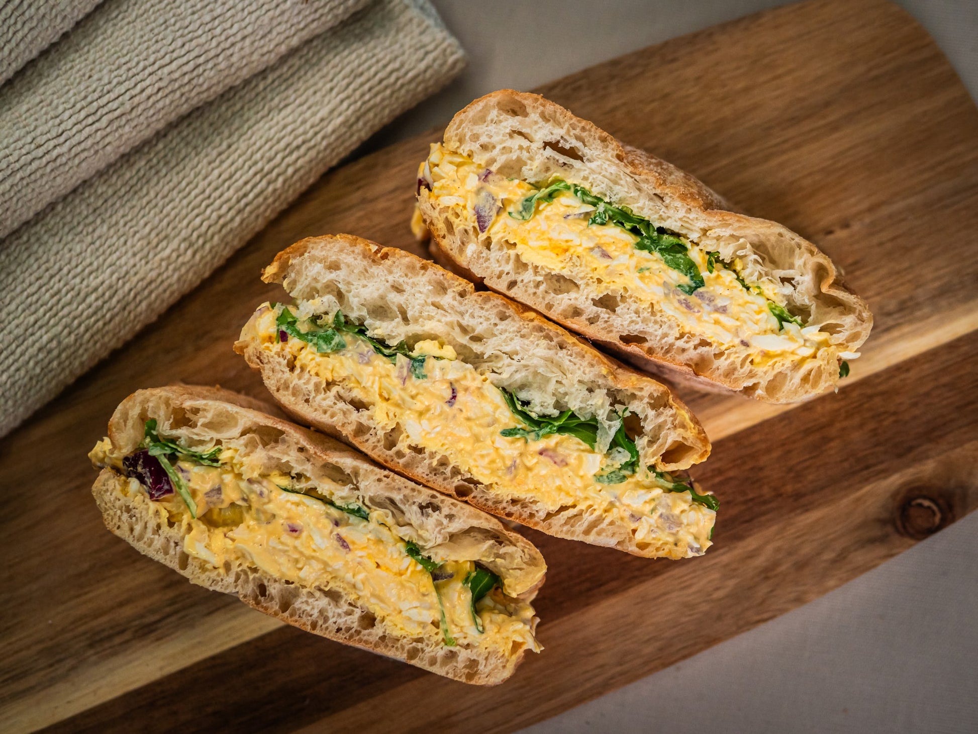 The Sunshine Stack by Sub Side, featuring three halves of sandwiches with lemon egg salad and fresh greens, is served on a wooden board beside folded beige napkins.