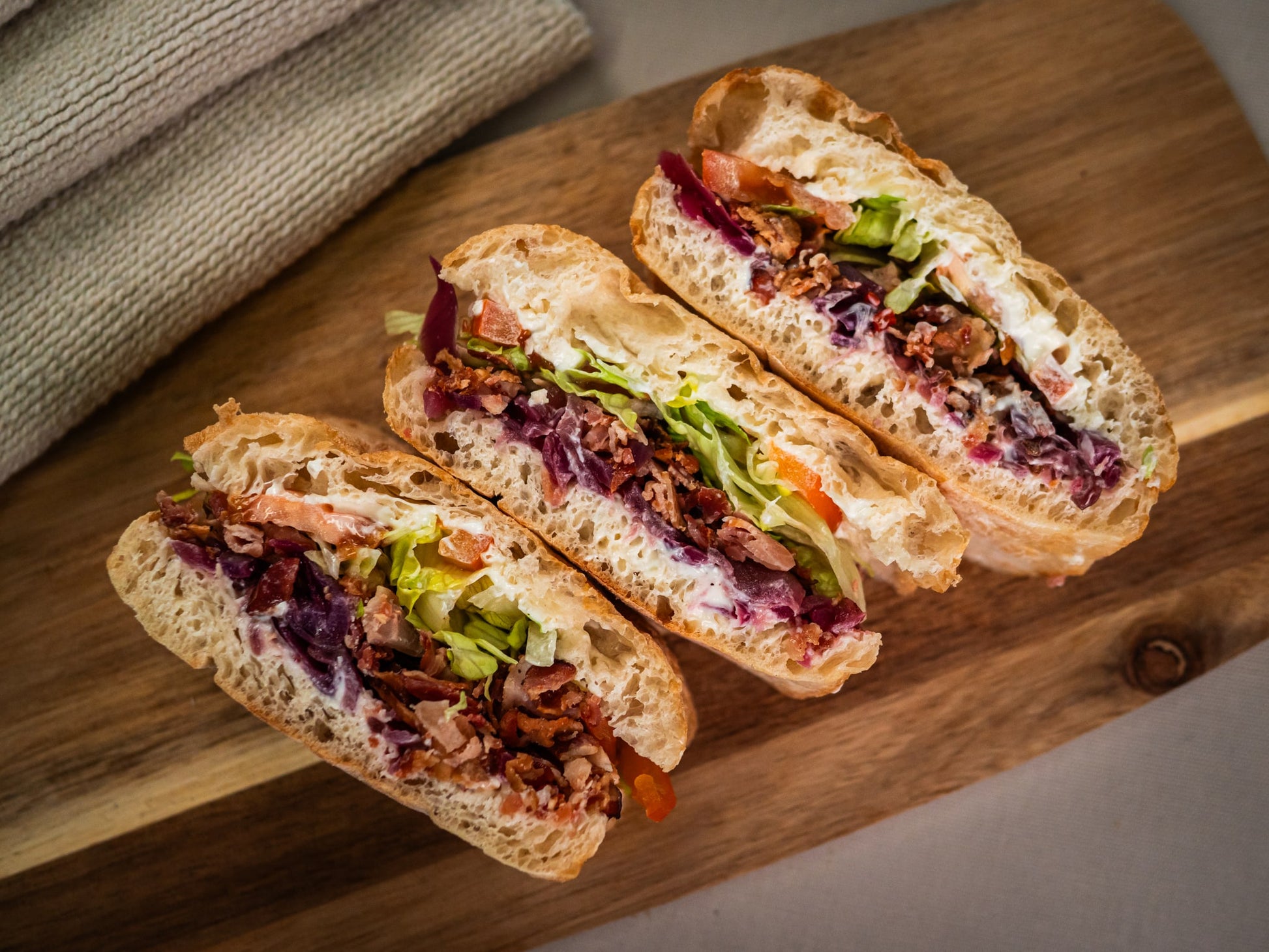 Three Blazin' Honey BLT sandwich halves by Sub Side, with crispy bacon, lettuce, tomato, creamy mayo, and hot honey red onions, are arranged on a wooden cutting board.