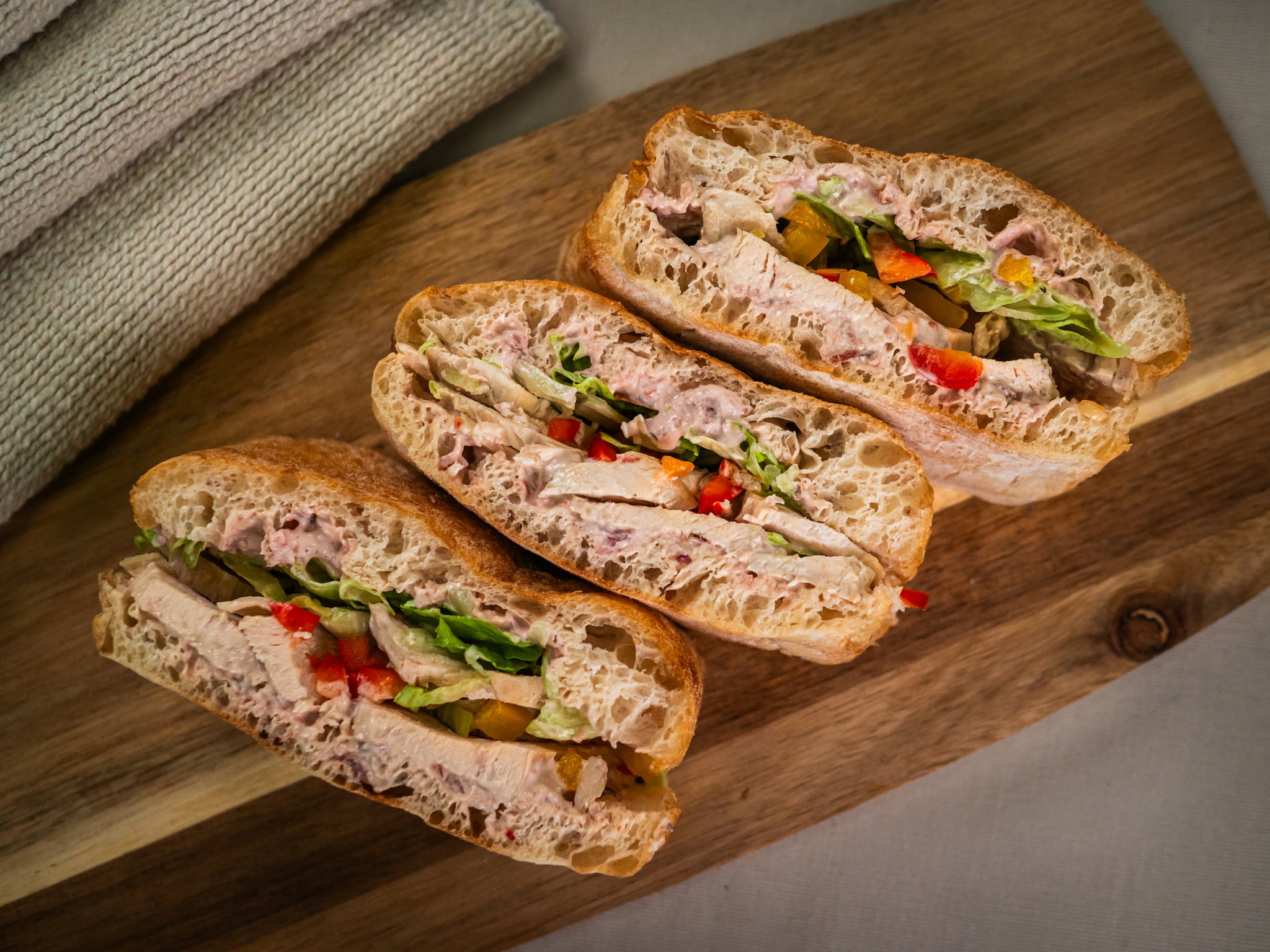 Three Sub Side Roasted Chicken Crunch ciabatta sandwiches with sliced chicken breast, lettuce, mixed peppers, and hot red onion mayo are arranged on a wooden serving board.