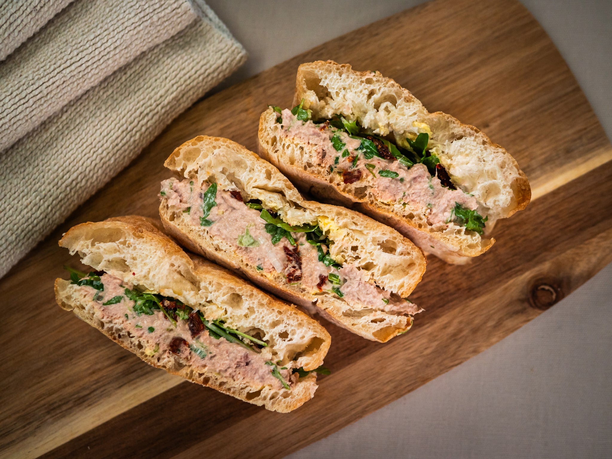 Three halves of Sub Side's Tuna Delight ciabatta sandwiches with lemon-Dijon tuna salad, peppery rocket, and greens are arranged on a wooden board beside folded napkins.