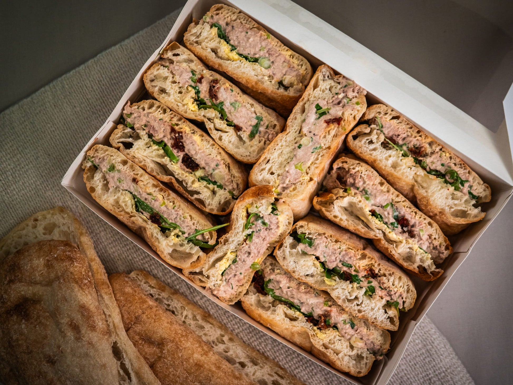 The Sub Side Tuna Delight features halved sandwiches filled with lemon-Dijon tuna, greens, and sun-dried tomatoes, neatly arranged beside pieces of rustic bread.