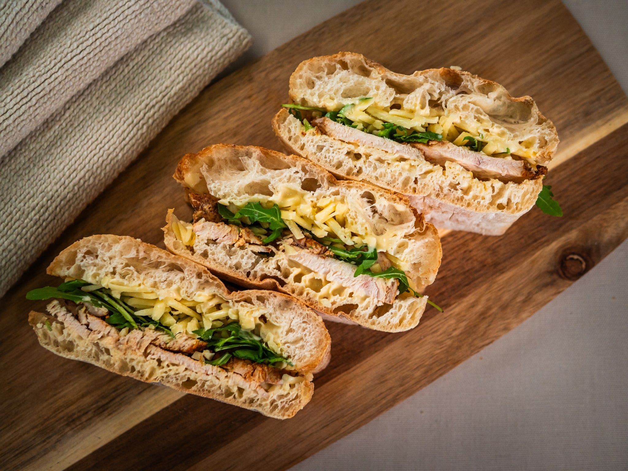 Three halves of Sub Side’s Zaátar Zing ciabatta sandwiches, filled with spiced chicken, cheese, and arugula, are arranged on a wooden cutting board.