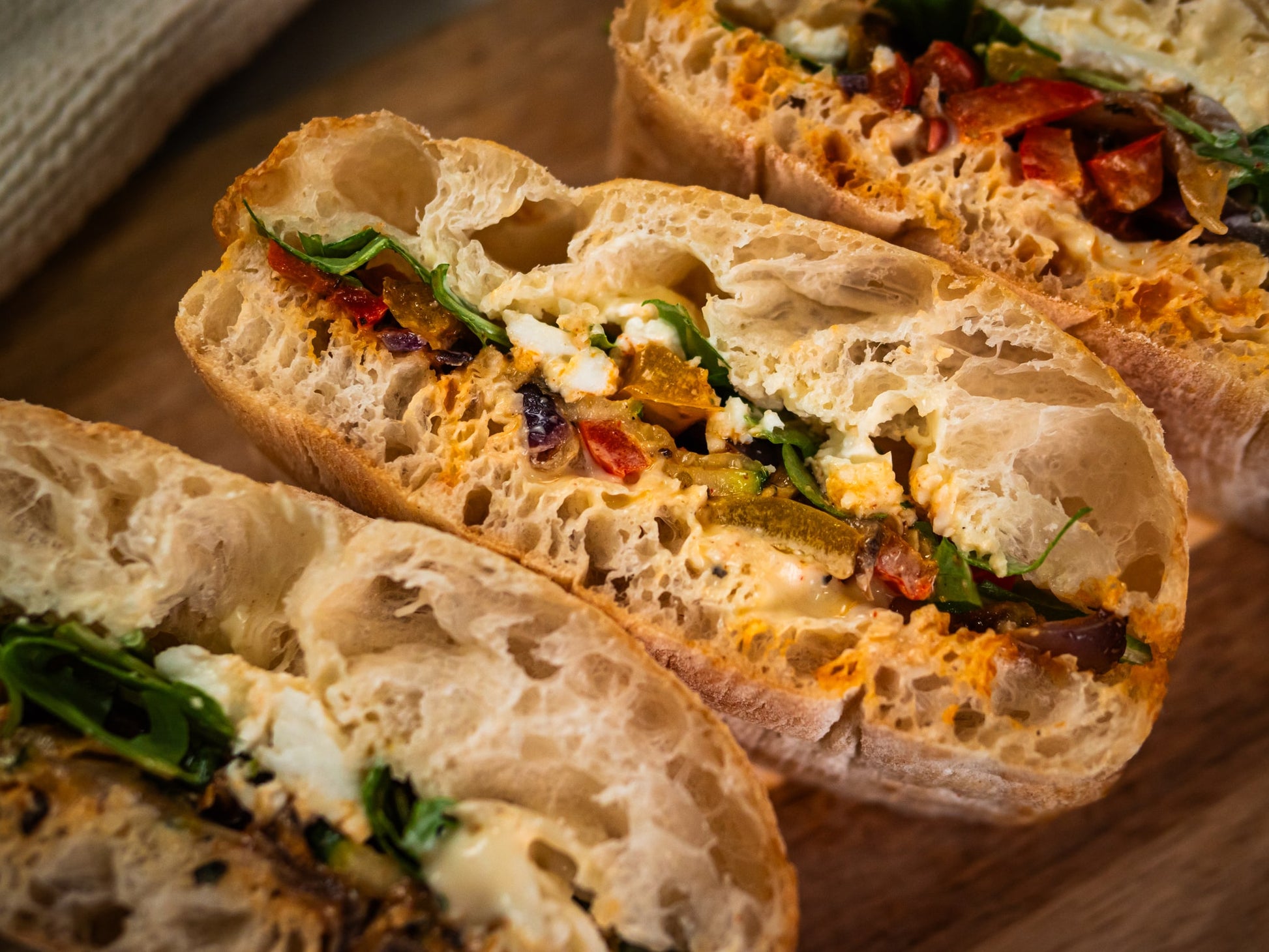 Sub Side’s Veggie Med features three crusty sandwich halves filled with mixed vegetables, greens, mozzarella cheese, and spreads, arranged on a wooden surface for a tasty vegetarian treat.