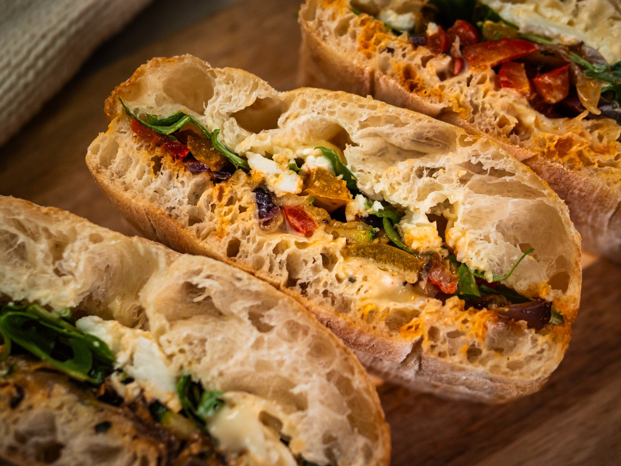 Sub Side’s Veggie Med features three crusty sandwich halves filled with mixed vegetables, greens, mozzarella cheese, and spreads, arranged on a wooden surface for a tasty vegetarian treat.