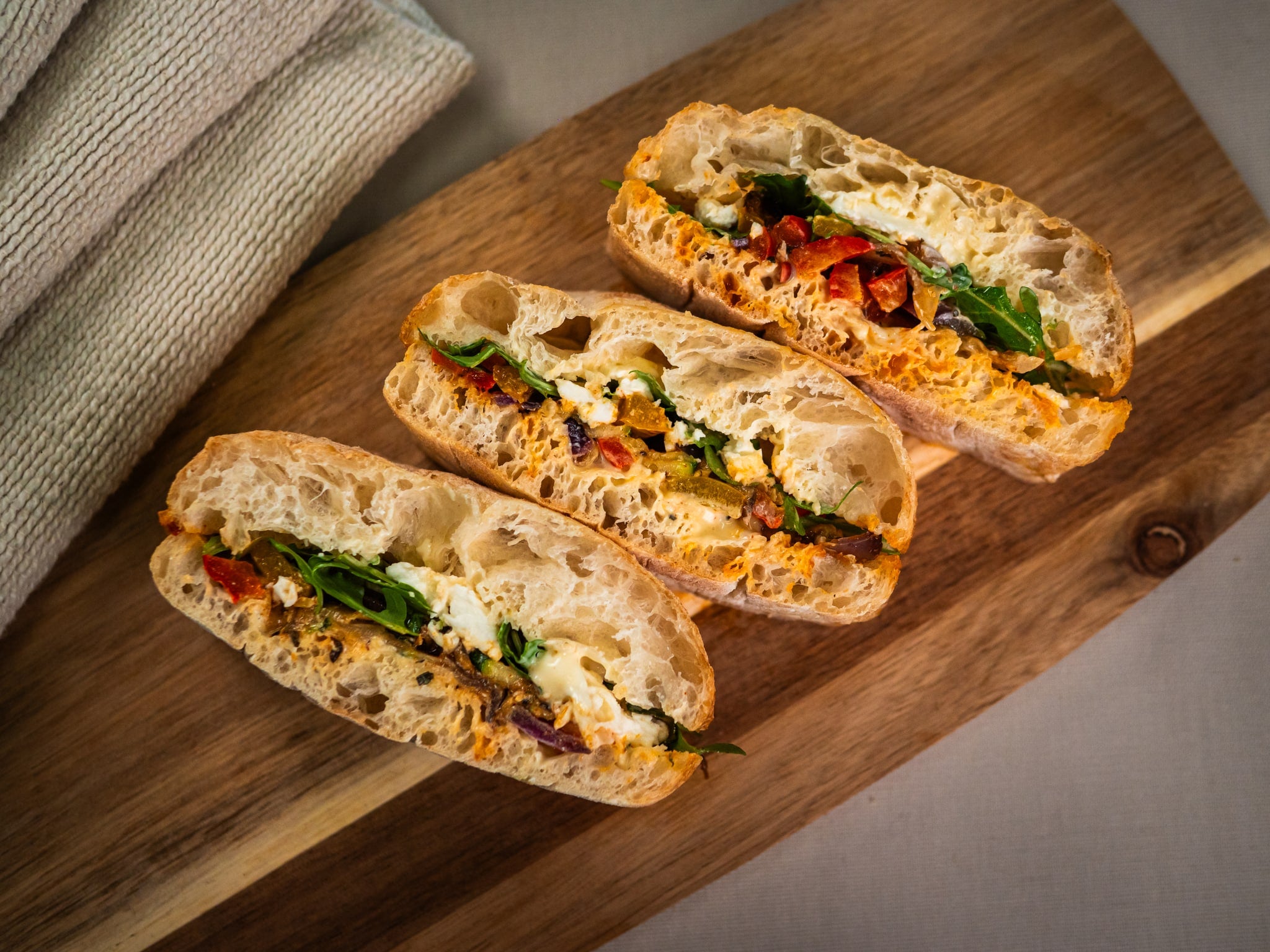 Three ciabatta sandwiches with greens, cheese, and roasted vegetables are arranged on a wooden board next to a folded beige cloth.
