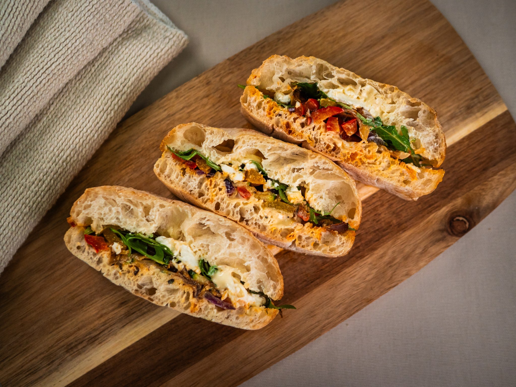 Three Sub Side Veggie Med ciabatta sandwiches with greens, roasted peppers, and mozzarella are arranged on a wooden board beside a folded cloth.
