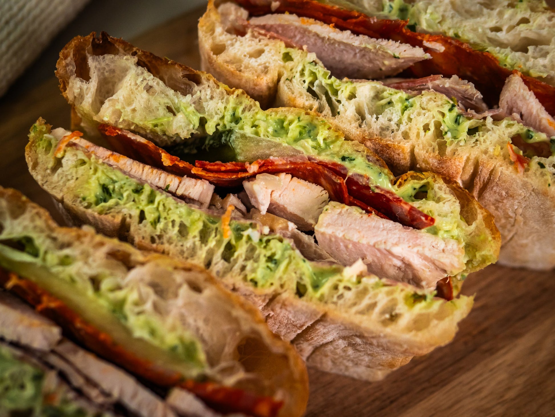 Close-up of the Hearty Sub Side by Sub Side: crusty bread sliced and layered with lettuce, roasted chicken, cheese, and creamy pesto mayo, arranged on a wooden surface.