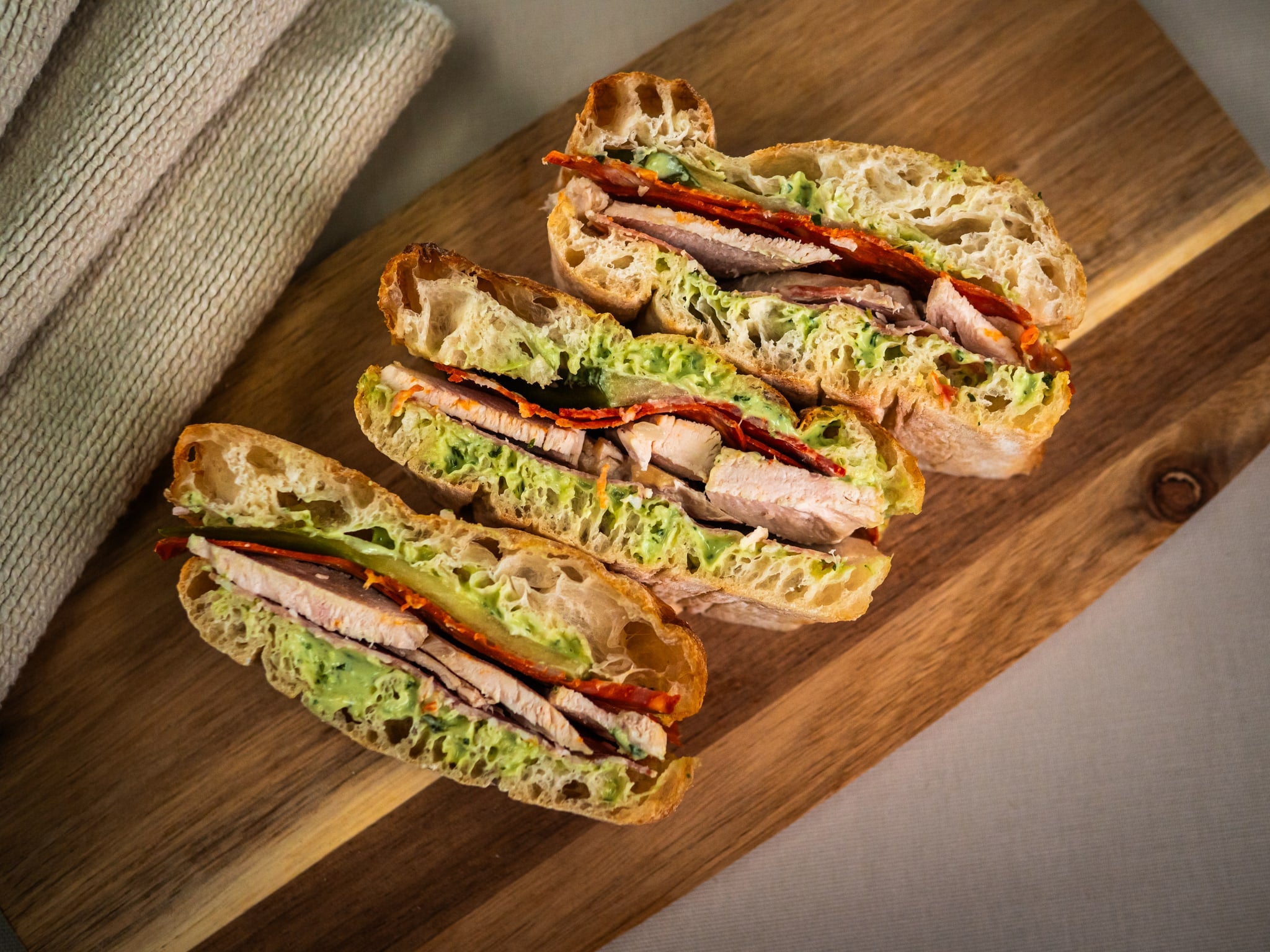Three halves of the Hearty Sub Side by Sub Side, featuring roasted chicken, lettuce, tomato, and creamy pesto mayo on ciabatta, are arranged on a wooden board with a folded cloth—a Mediterranean feast awaits.