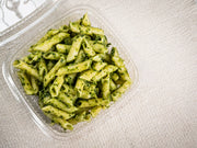 A clear plastic container of Sub Side Nut Free Pesto Pasta (500g) sits on a light textured surface, showcasing penne coated in fresh basil pesto sauce.