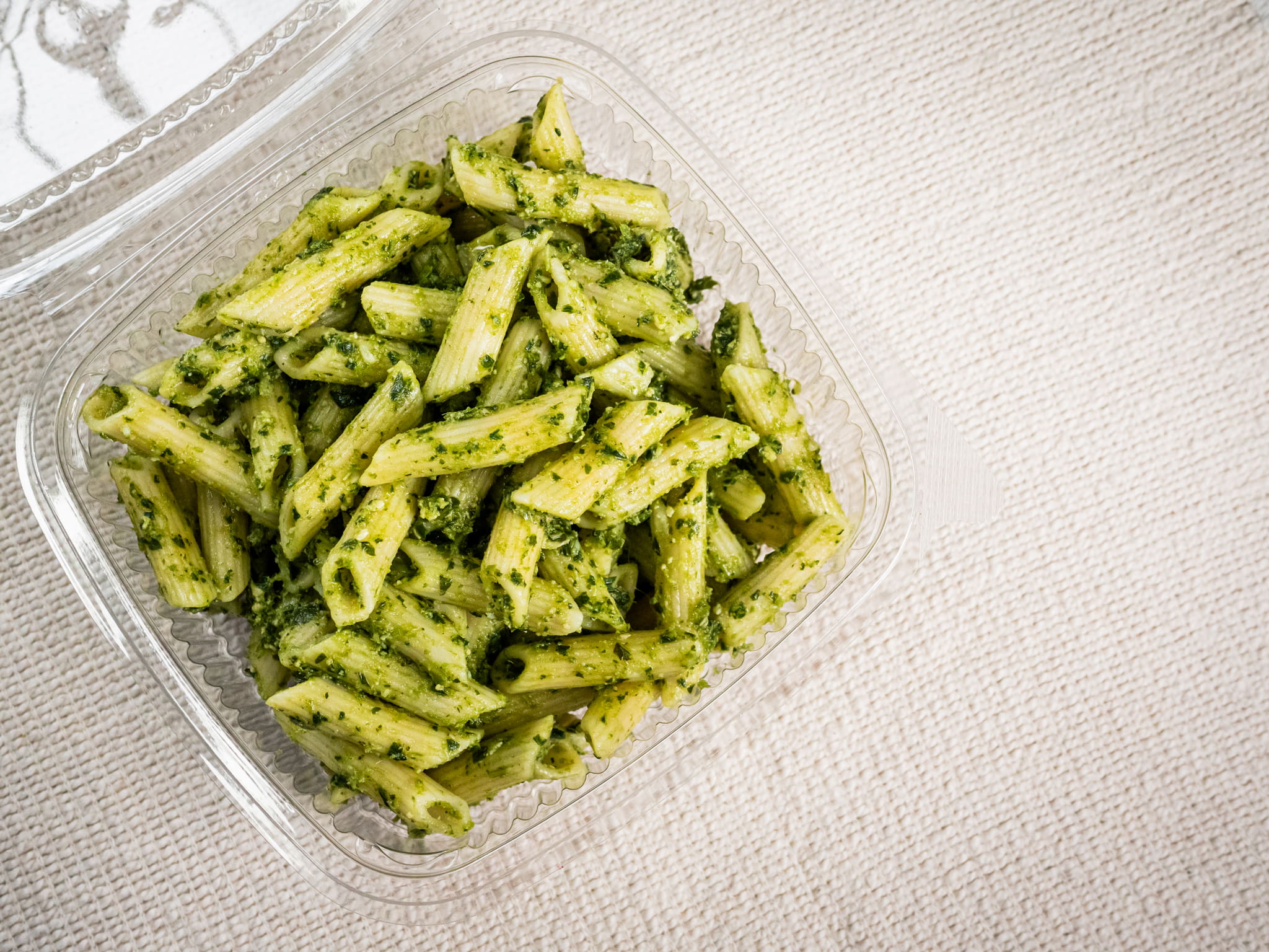 A clear plastic container of Sub Side Nut Free Pesto Pasta (500g) sits on a light textured surface, showcasing penne coated in fresh basil pesto sauce.