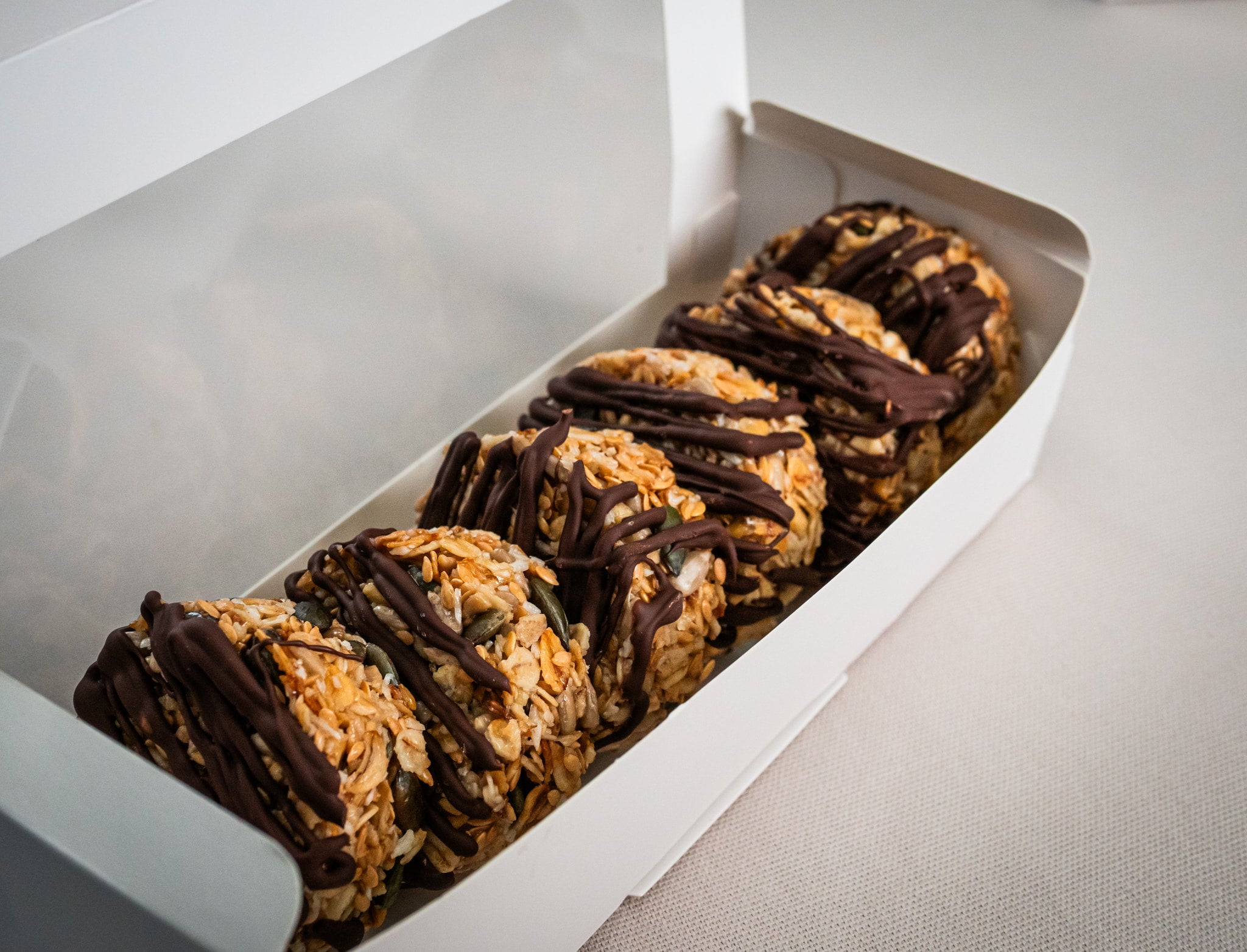 Six Sub Side Granola Bites Dark Chocolate, made with roasted oats and seeds and drizzled with dark chocolate, are arranged in a white box on a light surface—an ideal natural energy boost.