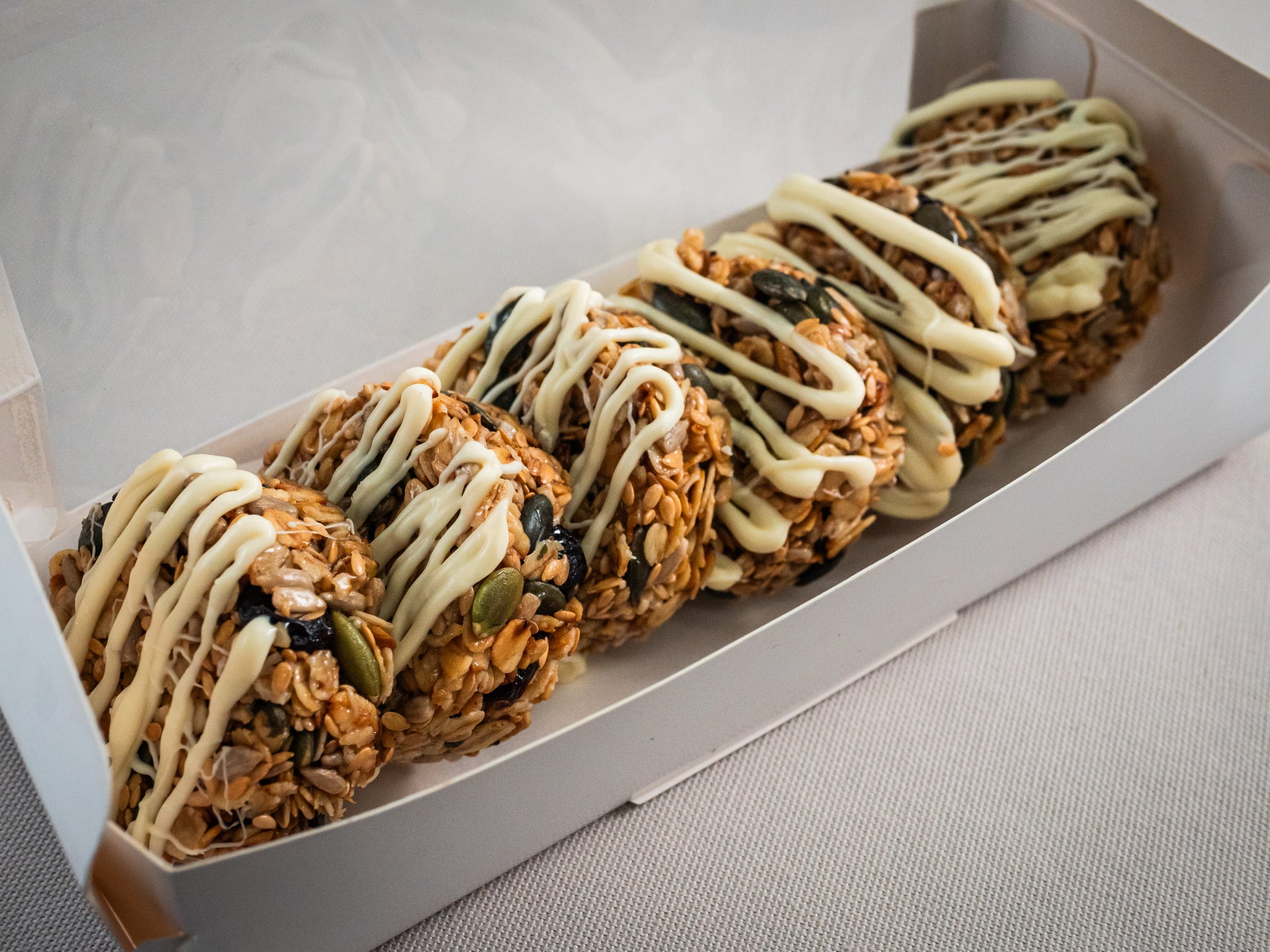 Six Sub Side Granola Bites White Chocolate, featuring house-roasted oats and dried cranberries with a white chocolate drizzle, are presented in a neat row inside a white rectangular box.