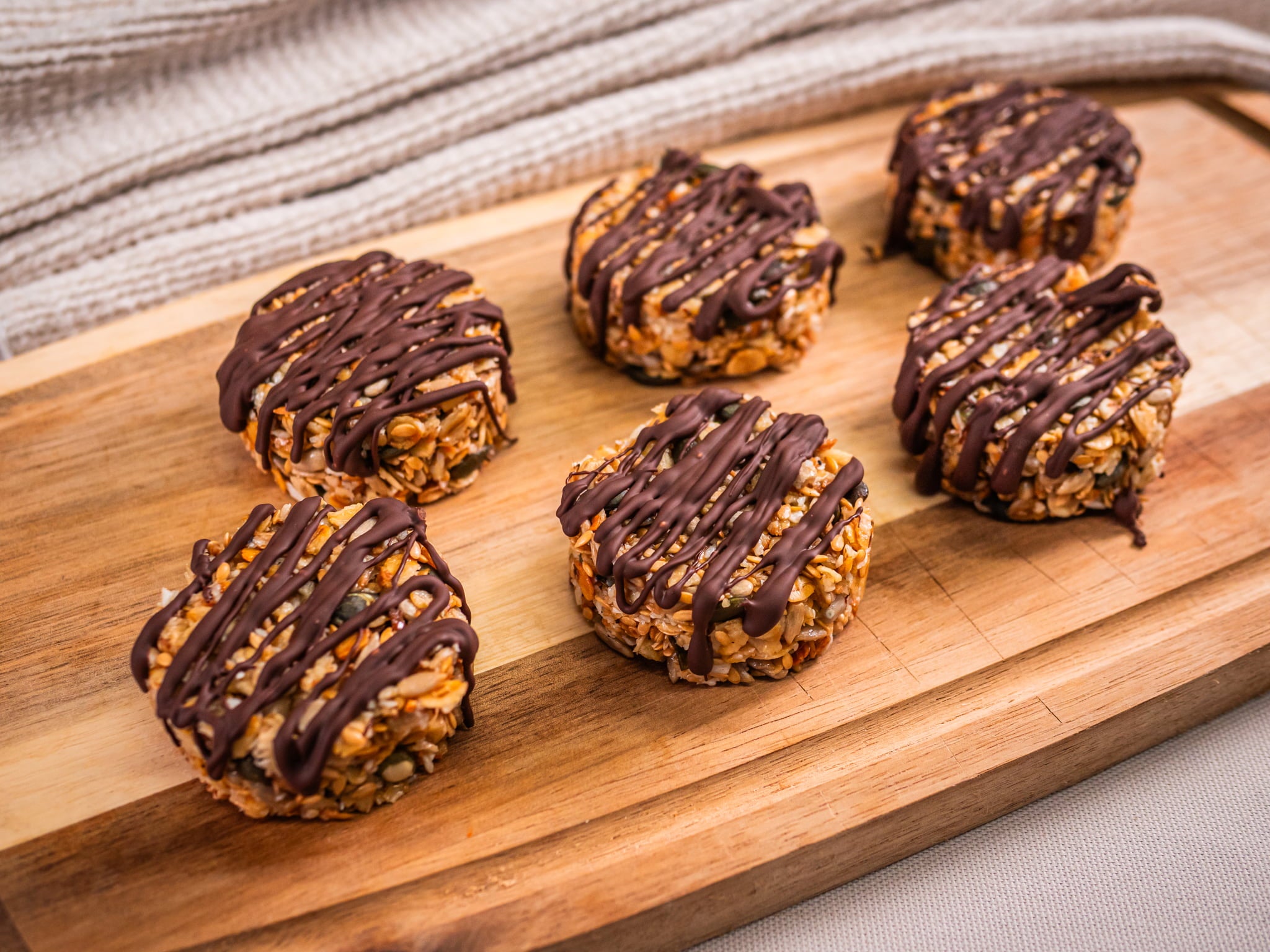 Sub Side’s Granola Bites Dark Chocolate (6 Pieces), made with roasted oats and drizzled with dark chocolate, are arranged on a wooden cutting board with a textured cloth in the background.