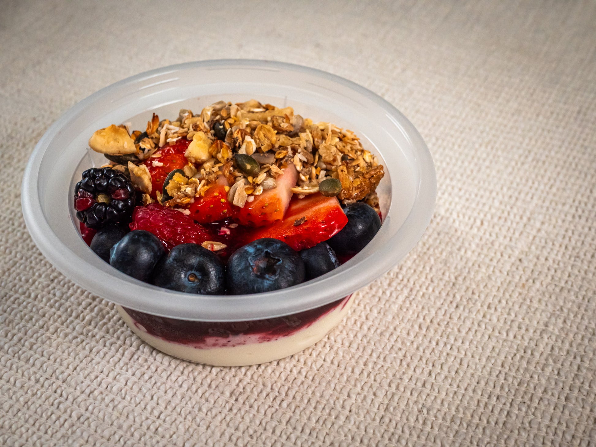The Sub Side Yogurt Berry Snap Pot (227g) features Greek yogurt with blueberries, strawberries, blackberries, homemade granola, and seeds in a plastic container on a textured beige surface.