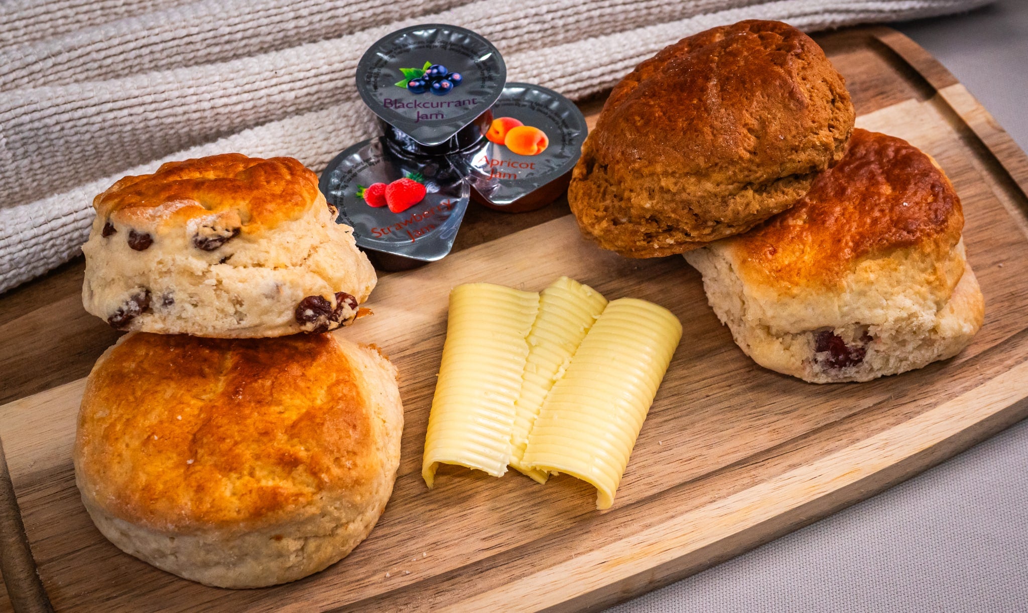 Sub Side’s Freshly Baked Scones (10 pieces) are served with butter curls and fruit jam portions on a wooden board, with a towel in the background.