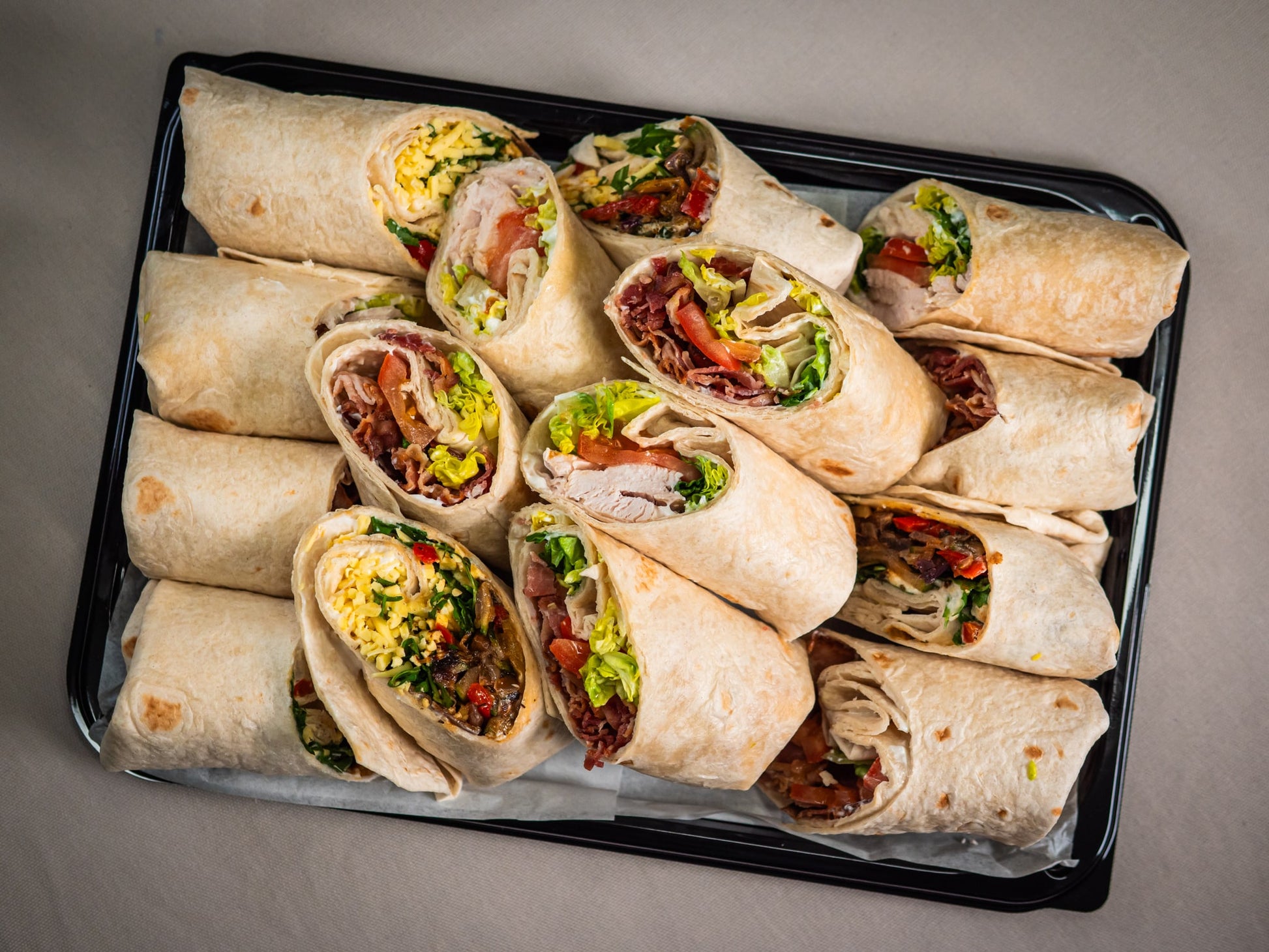 The Sub Side Mixed Wrap Platter (serves 7) features a black tray filled with an assortment of colorful tortilla wraps, each cut open to showcase fillings like lettuce, scrambled eggs, sliced meats, and fresh vegetables.