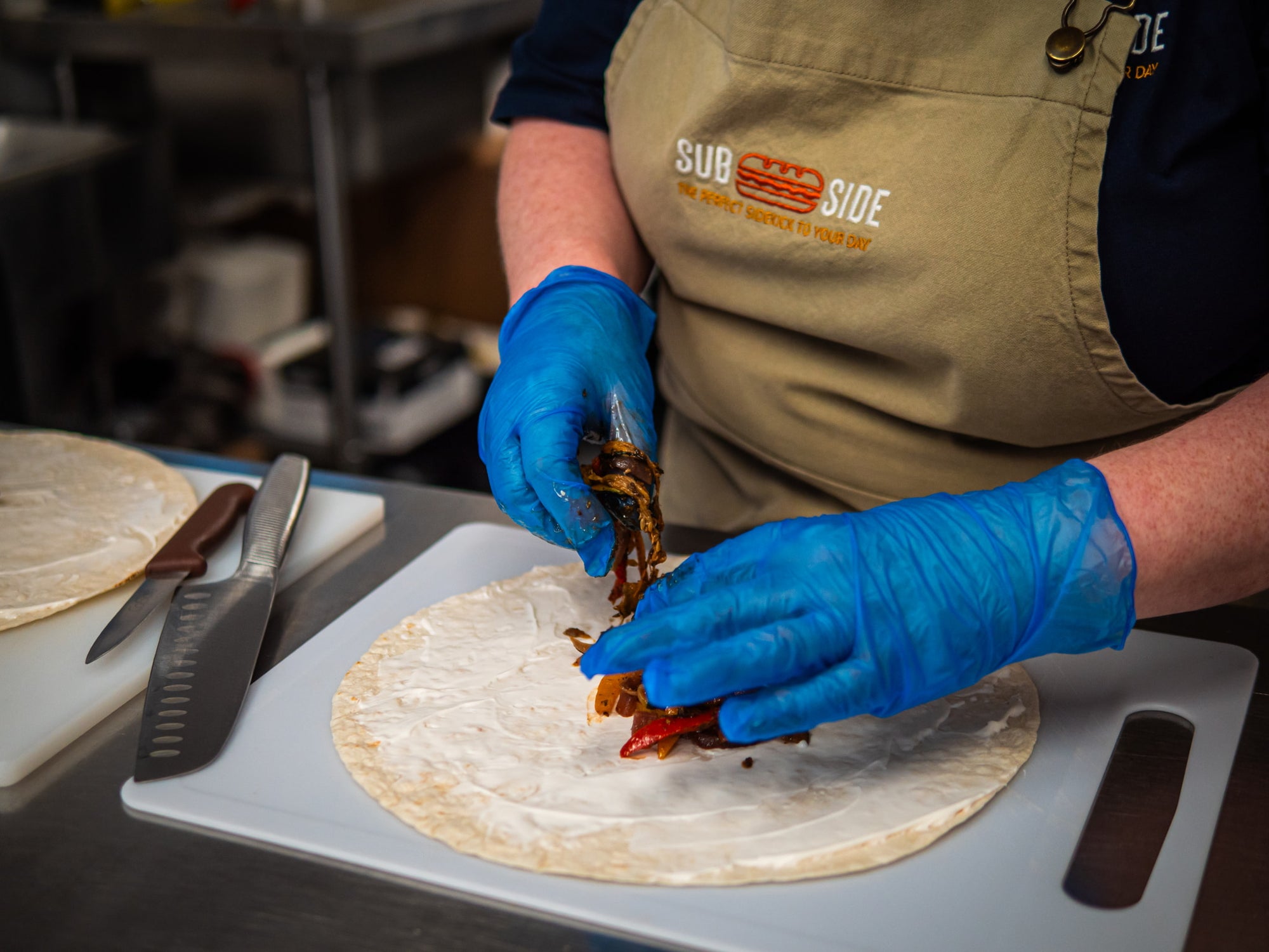 A person wearing blue gloves and a tan apron prepares a wrap by adding cooked vegetables onto a tortilla on a cutting board in a kitchen.