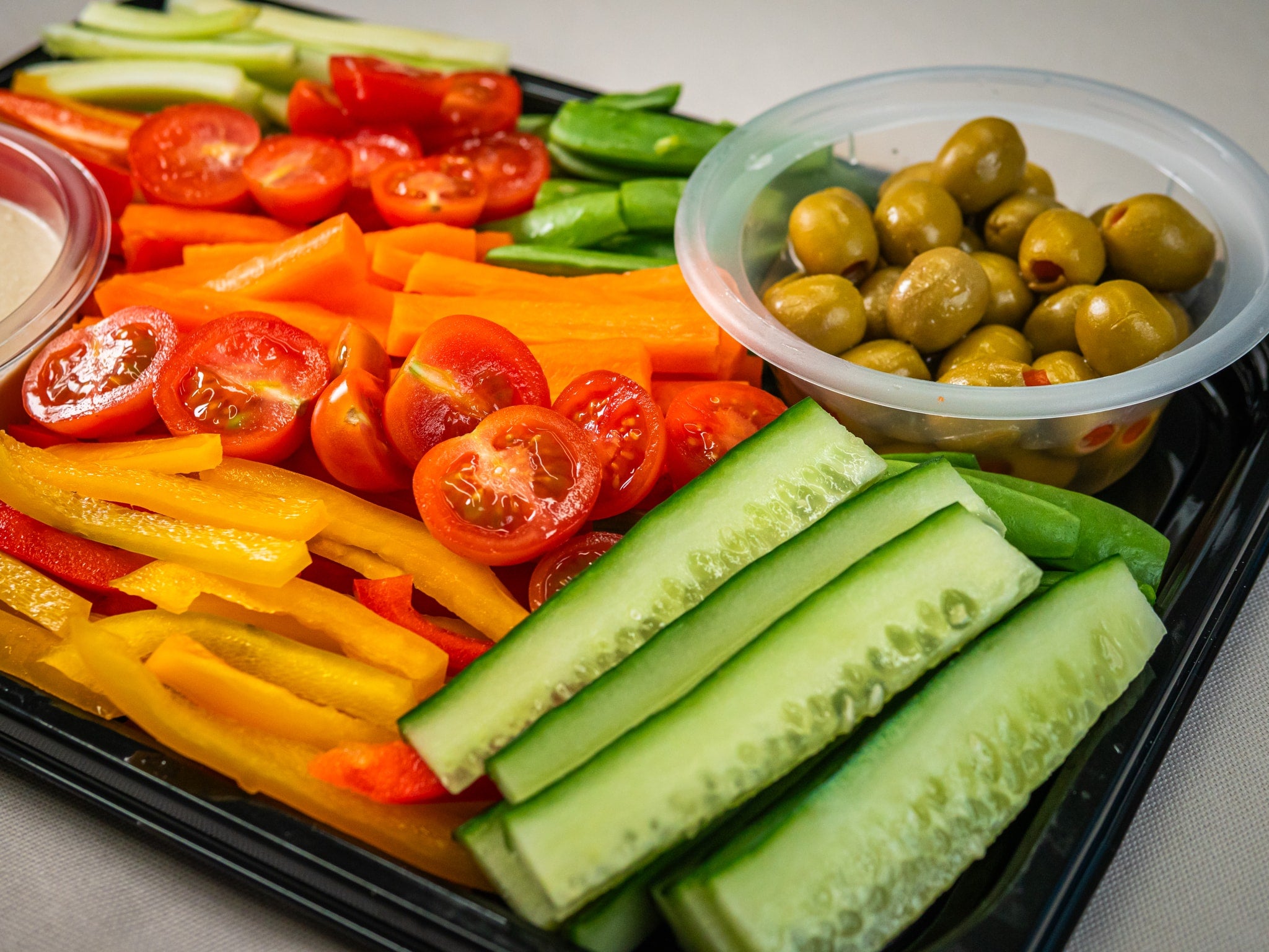 A tray with sliced cucumbers, bell peppers, cherry tomatoes, snap peas, green olives in a plastic container, and a portion of hummus.
