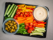 The Sub Side Garden Fresh Veggie Platter (serves 8 pax) features sliced cucumbers, bell peppers, carrots, celery, cherry tomatoes, snap peas, marinated olives, and creamy hummus on a beige surface.