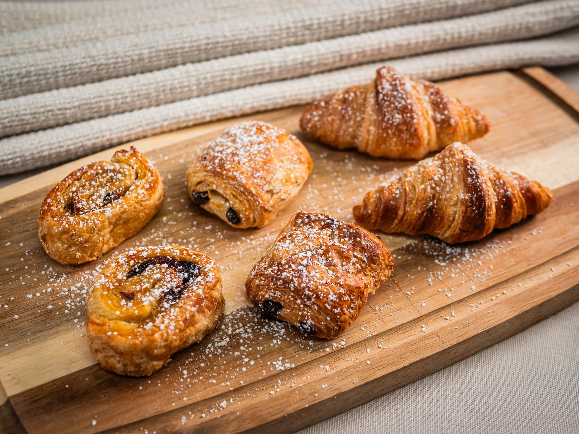 Sub Side’s Assorted Mini Pastries (22 pieces) feature butter croissants and pains au chocolat, all dusted with powdered sugar and elegantly arranged on a wooden board.
