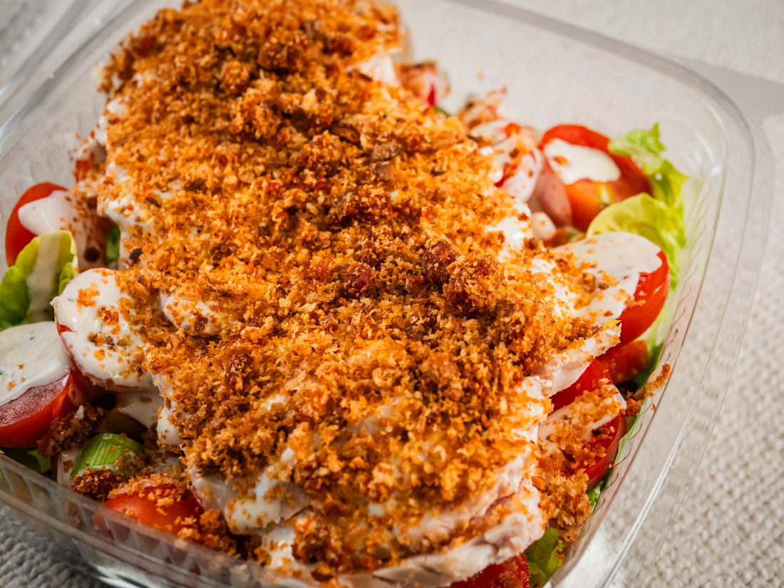 A clear plastic container filled with salad ingredients, topped with a generous layer of crispy golden brown breadcrumbs and creamy dressing.