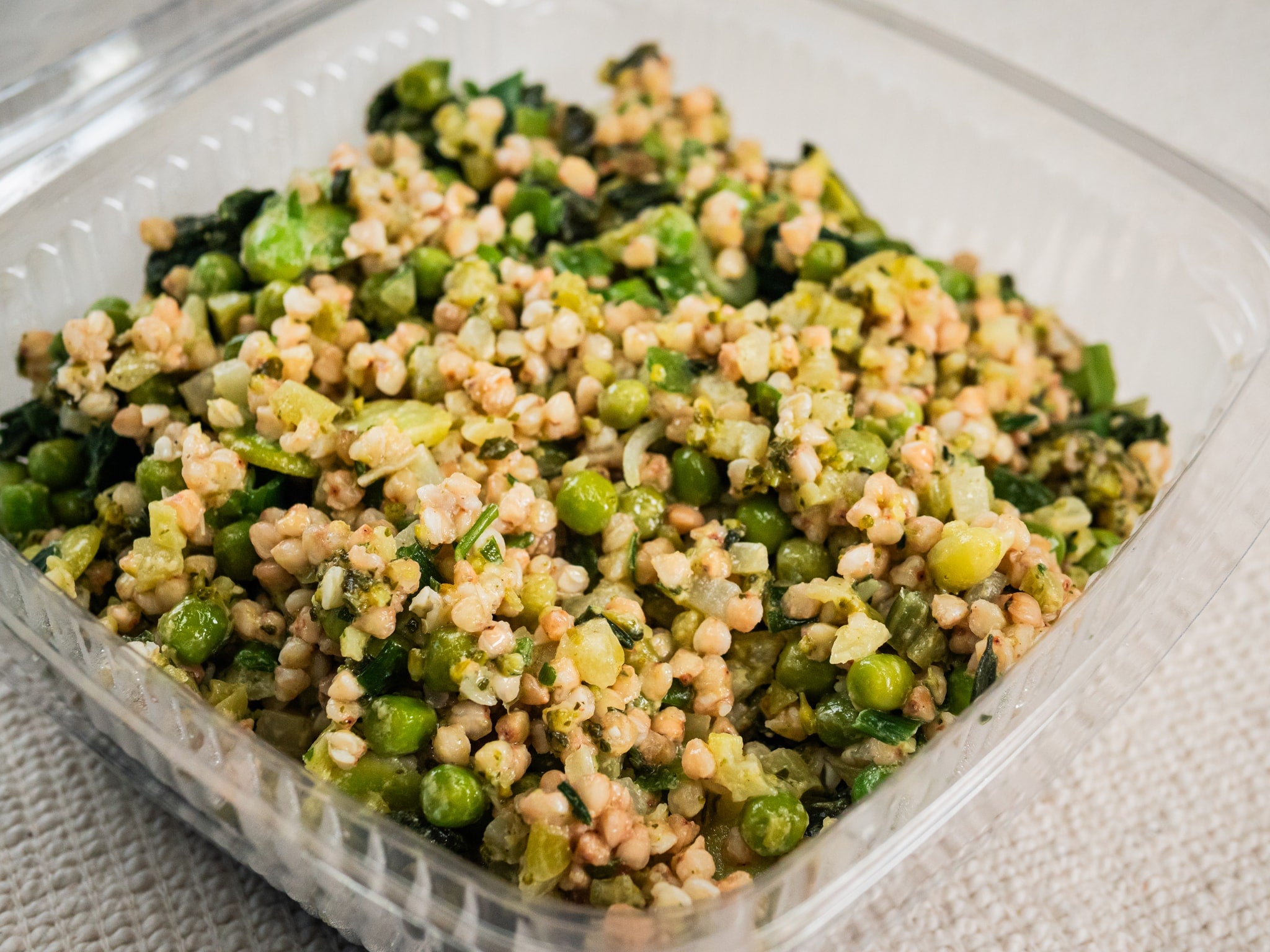 A 500g Sub Side Green Wheat Salad in a clear plastic container, featuring garden peas, buckwheat, chopped greens, and herbs, sits on a light textured surface.