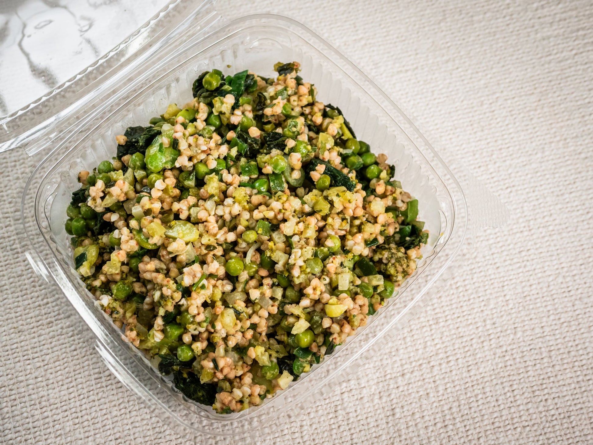 A Sub Side Green Wheat Salad (500g) in a clear plastic container, featuring buckwheat, garden peas, and leafy greens, rests on a textured light surface.