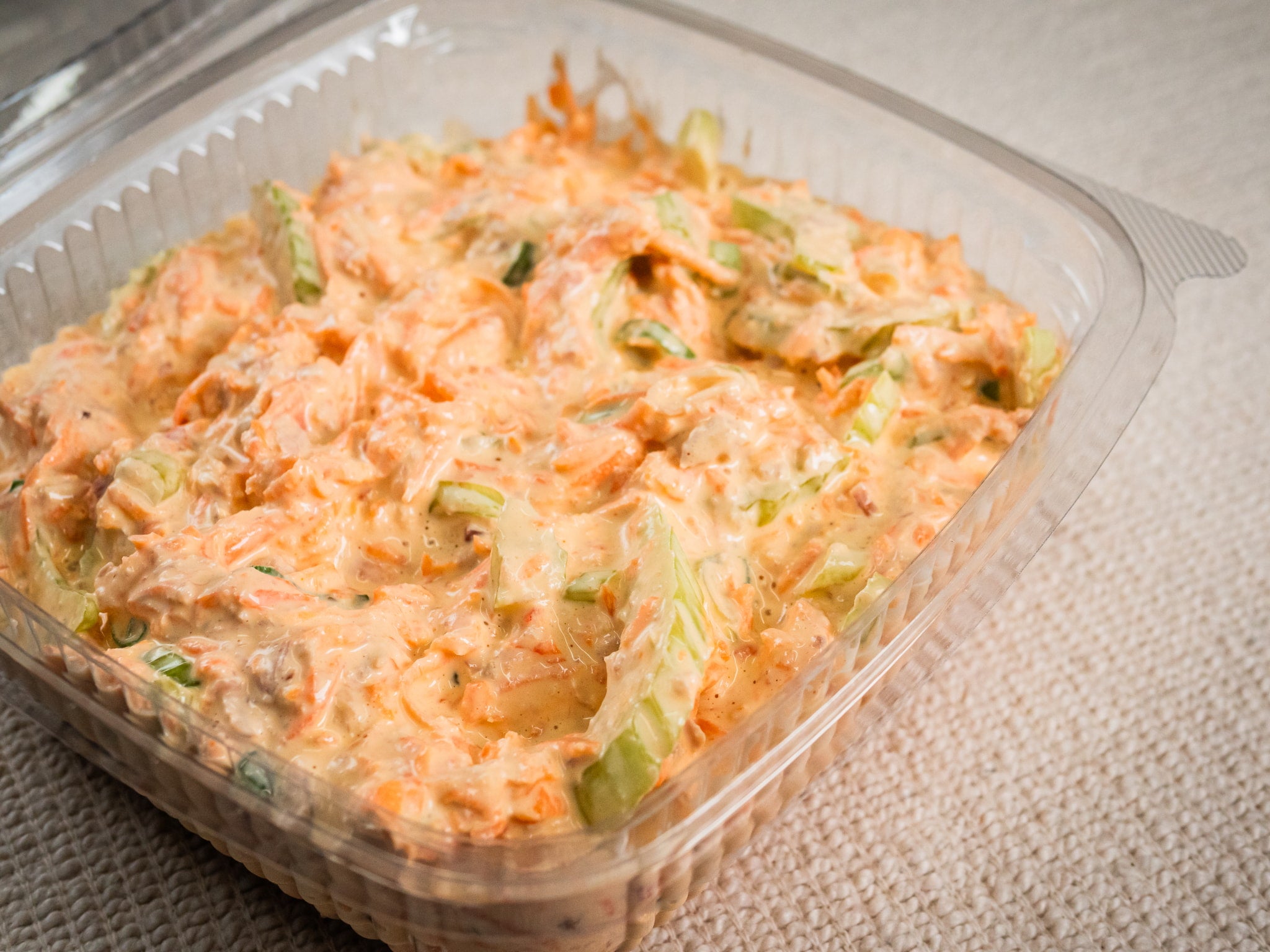 A clear plastic container of Sub Side Hot Honey Slaw, a creamy salad mix with crispy carrots, chopped celery, and fresh spring onions, sits on a textured surface.