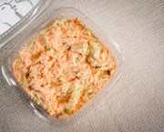 A clear plastic container of Sub Side's Hot Honey Slaw, a creamy shredded carrot and celery salad with crispy carrots and fresh spring onions, sits on a textured beige surface.