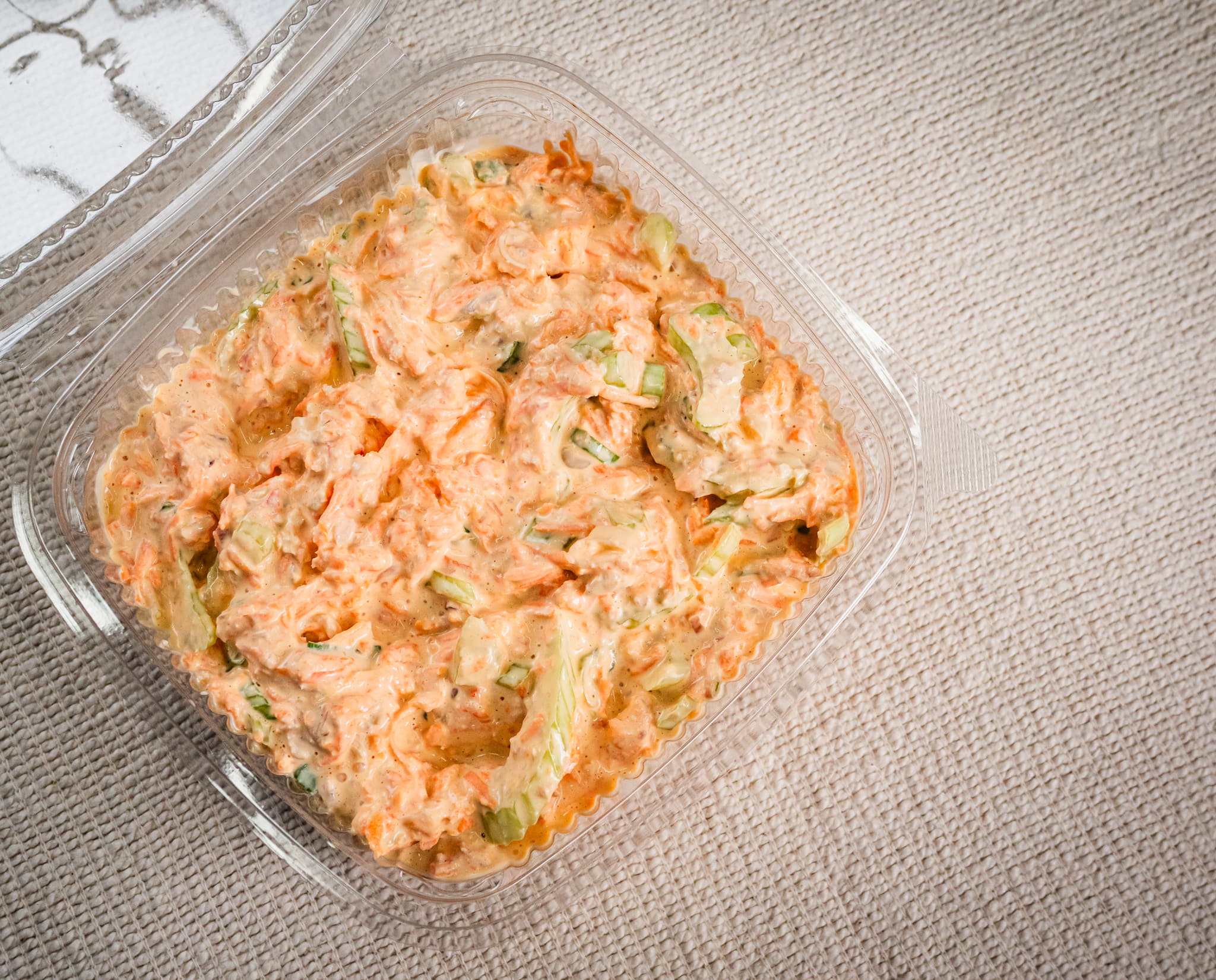 A clear plastic container of Sub Side's Hot Honey Slaw, a creamy shredded carrot and celery salad with crispy carrots and fresh spring onions, sits on a textured beige surface.
