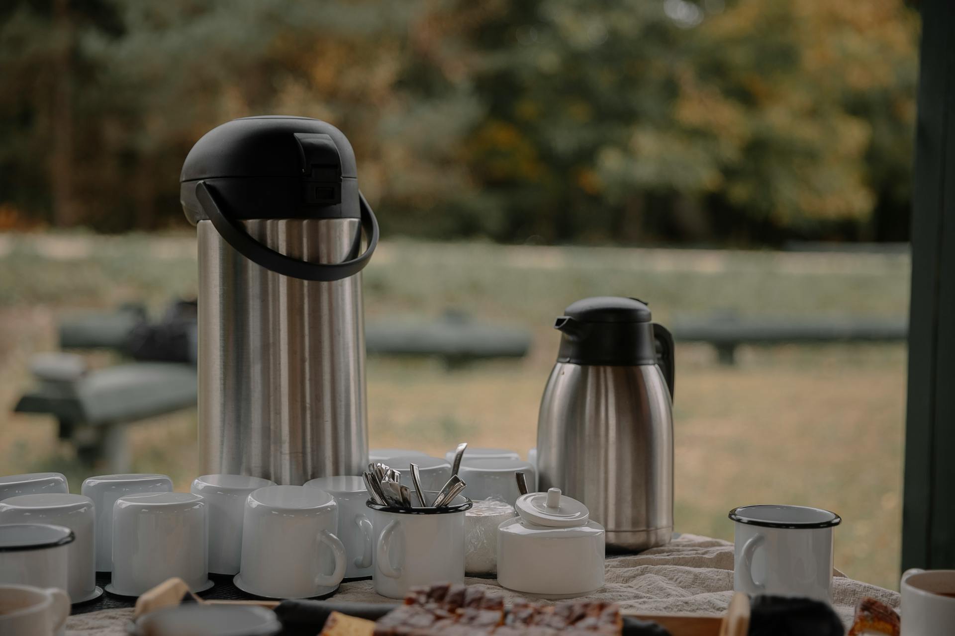 The Sub Side Tea & Coffee Service (20 pax) features large and small stainless steel coffee carafes, fresh filter coffee, white cups, sugar bowl, utensils, and pastries set outdoors with blurred trees in the background.
